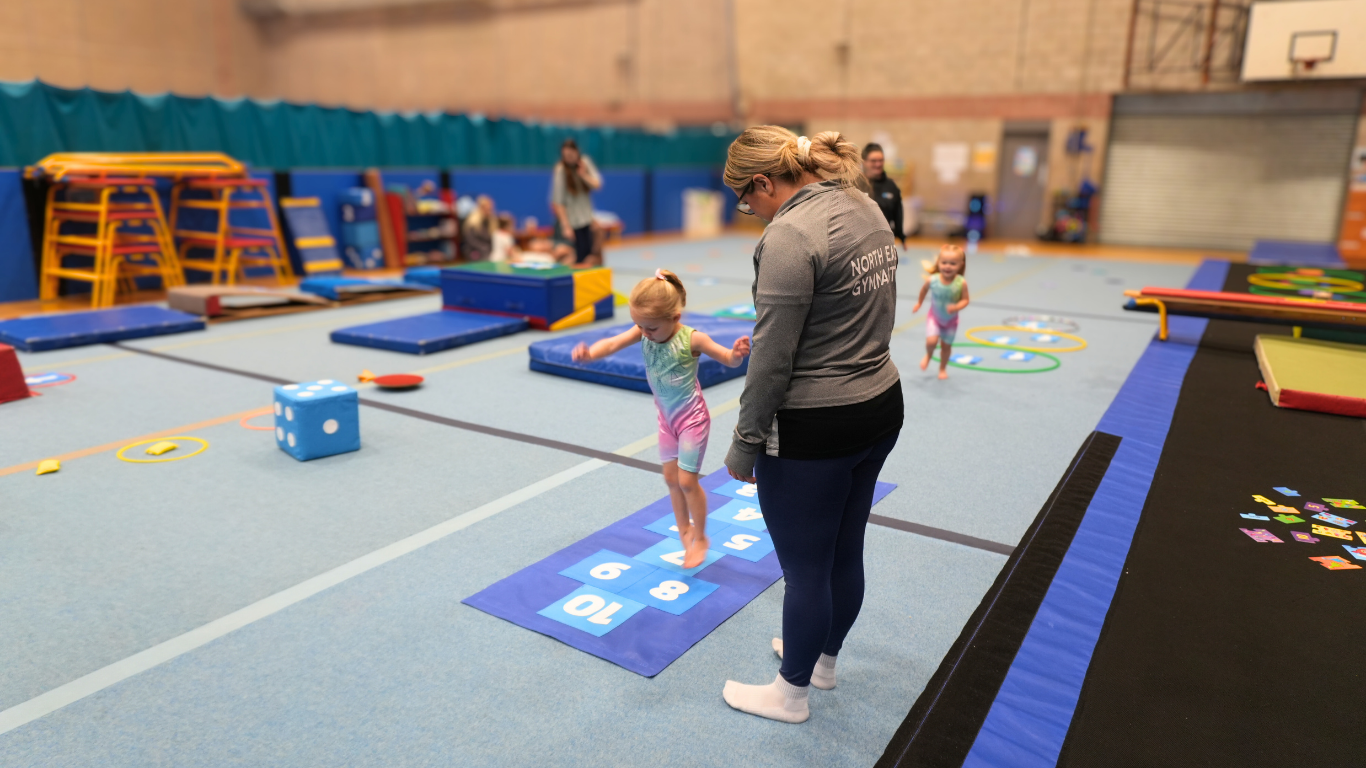 pre-school gymnast doing hop scotch with a coach watching closely